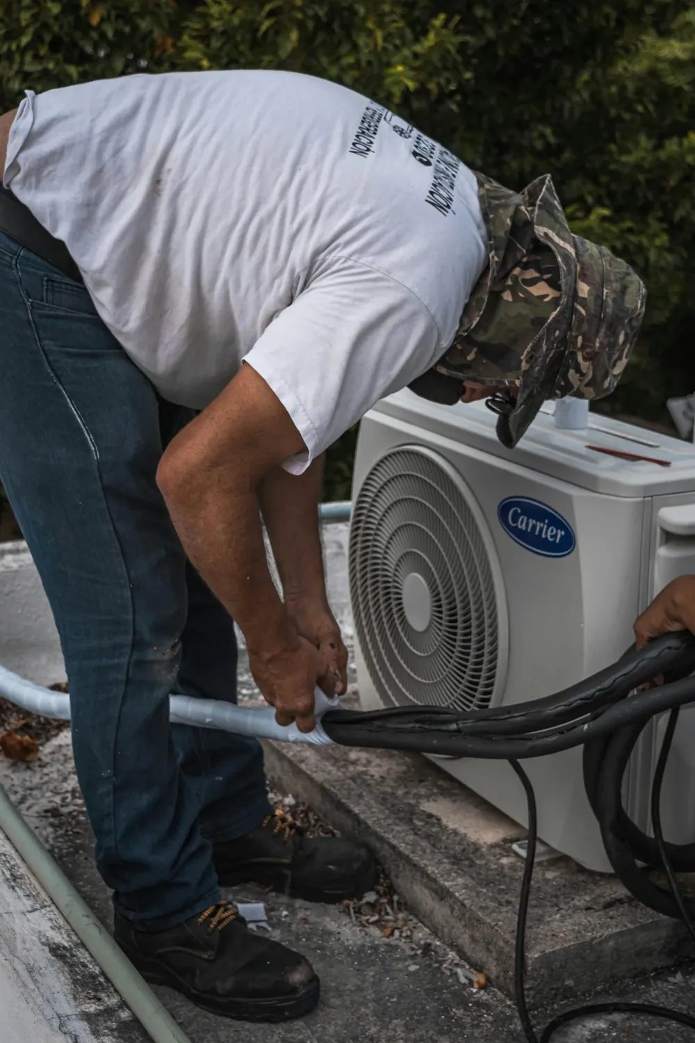 Technician working beside a Carrier outdoor condenser while handling insulated lines during installation. The photo highlights professional Air Conditioning Services for a residential system.