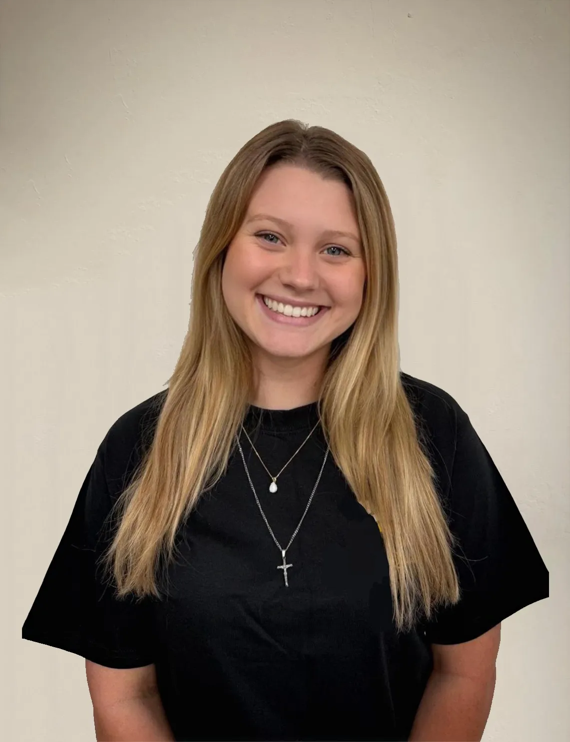 Isabella Gardner-Black smiling in a black shirt with layered necklaces, standing against a light neutral wall. Portrait of the Scheduling Specialist.
