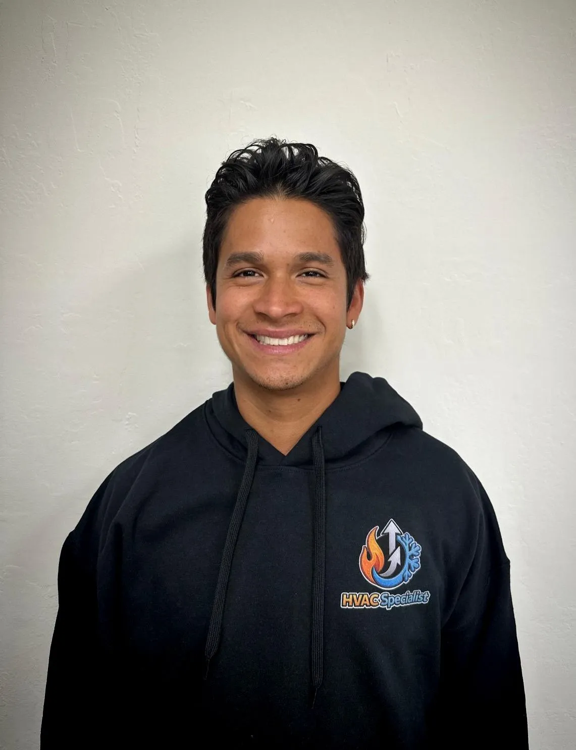 Luis Morales smiling in a black hoodie with the text "HVAC Specialist" and company logo on the front, standing against a light neutral wall. Portrait of an HVAC Specialist.