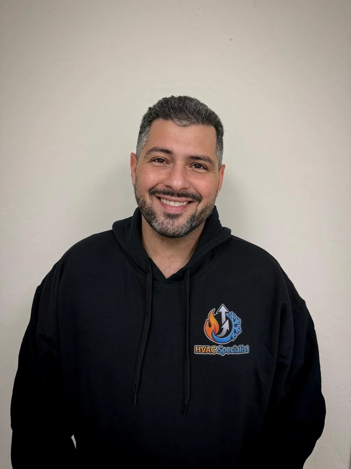 Sam Levi smiling in a black hoodie with the text "HVAC Specialist" and company logo on the front, standing against a light neutral wall. Portrait of the Lead HVAC Specialist.