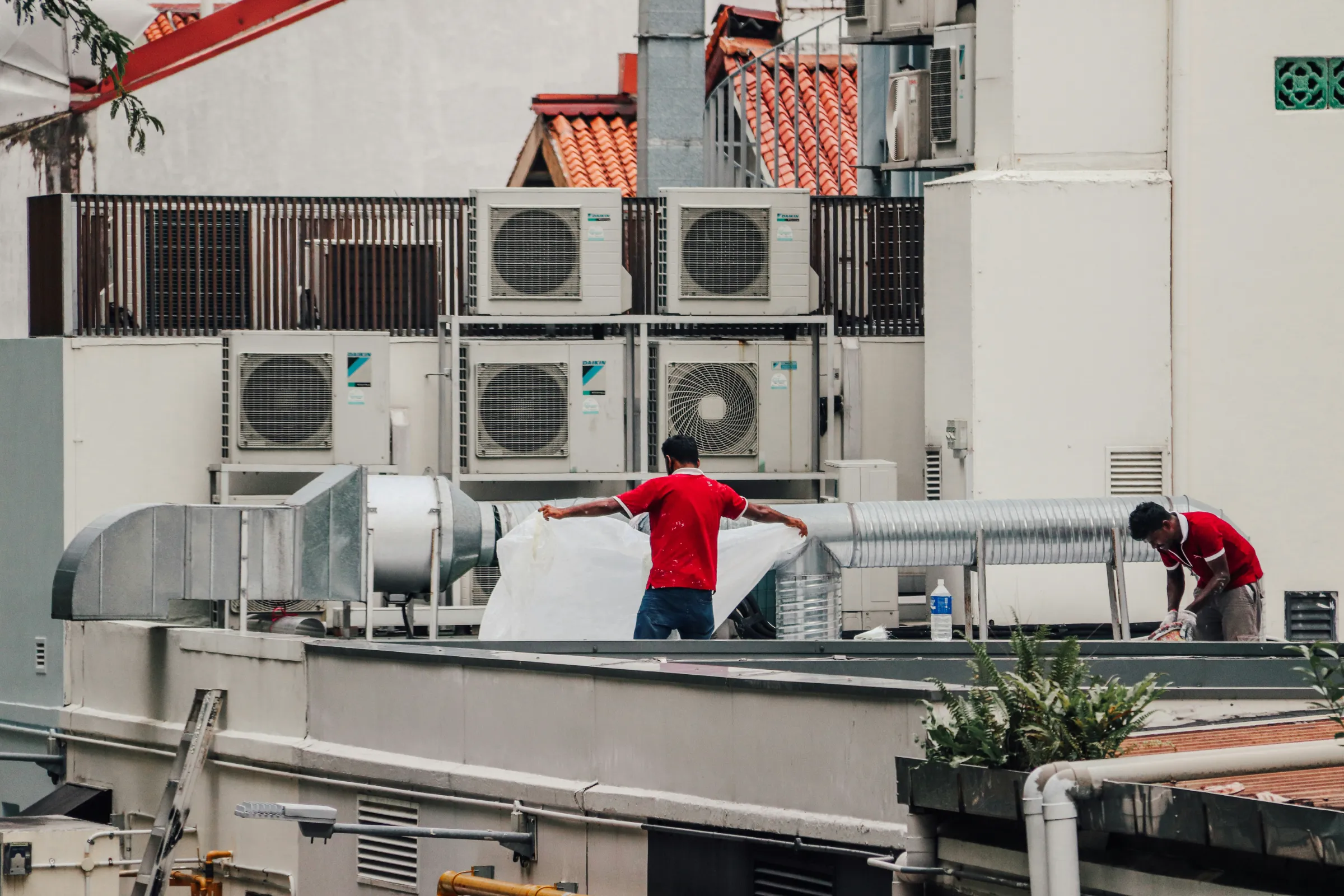Two technicians in red uniforms work on a commercial rooftop, handling a large flexible duct next to a metal ventilation duct, surrounded by multiple Daikin outdoor AC condenser units mounted on tiered platforms.